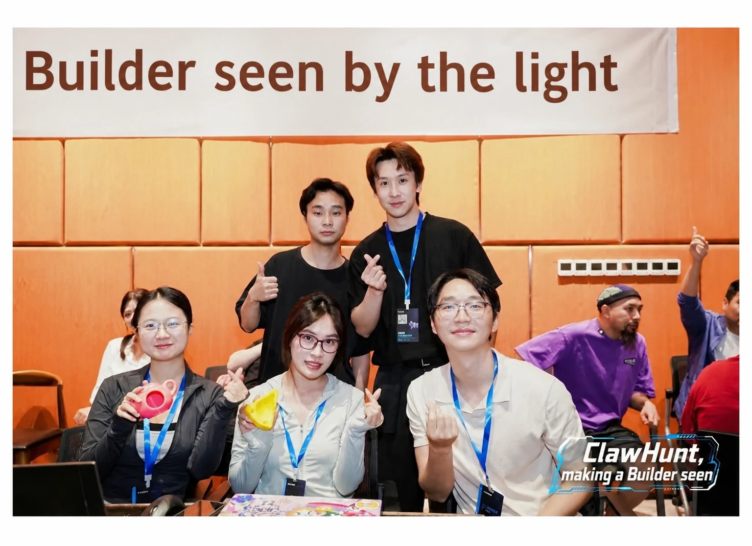 图片展示了一群人在室内合影的场景。背景墙上有“Builder seen by the light”字样。前排坐着五人,其中三人面带微笑,做出比心手势,均佩戴有挂绳。后排有三人在竖起大拇指,其中一人穿着黑色上衣,另一人穿着黑色T恤,还有一人穿着紫色上衣。画面右下角有“ClawHunt, making a Builder seen”的标志。此图与文档中介绍团队在一次72小时高强度比赛中,因网络不稳定等原因差点崩溃,但最终因一位投资者发现项目而获得投资的内容相关,体现了团队在比赛中的努力与坚持。